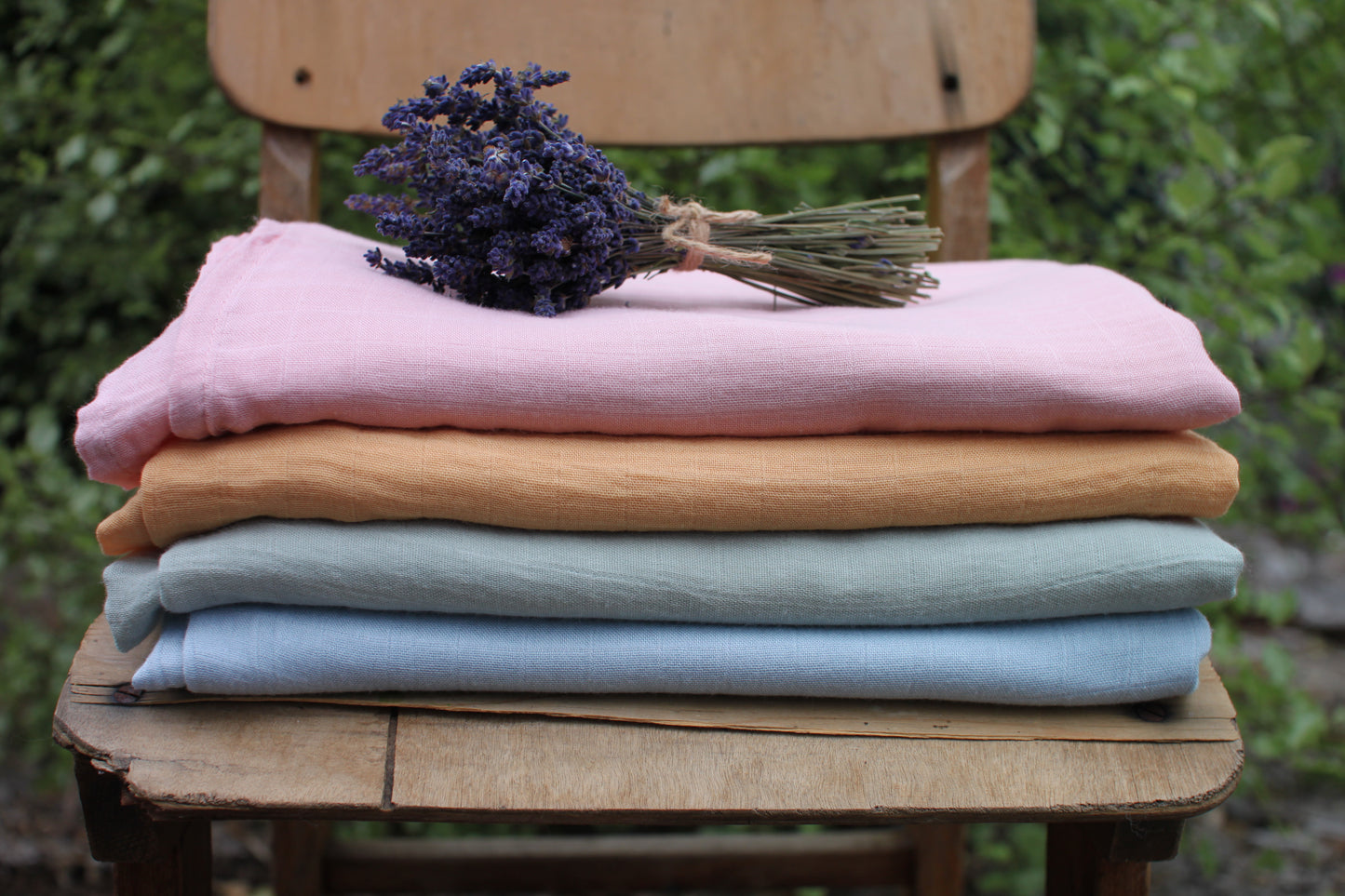Four muslin cloths are folded and stacked on a wooden chair. A bunch of dried lavender sits on top. The colours from top to bottom are pale pink, orange, moss green and sky blue.