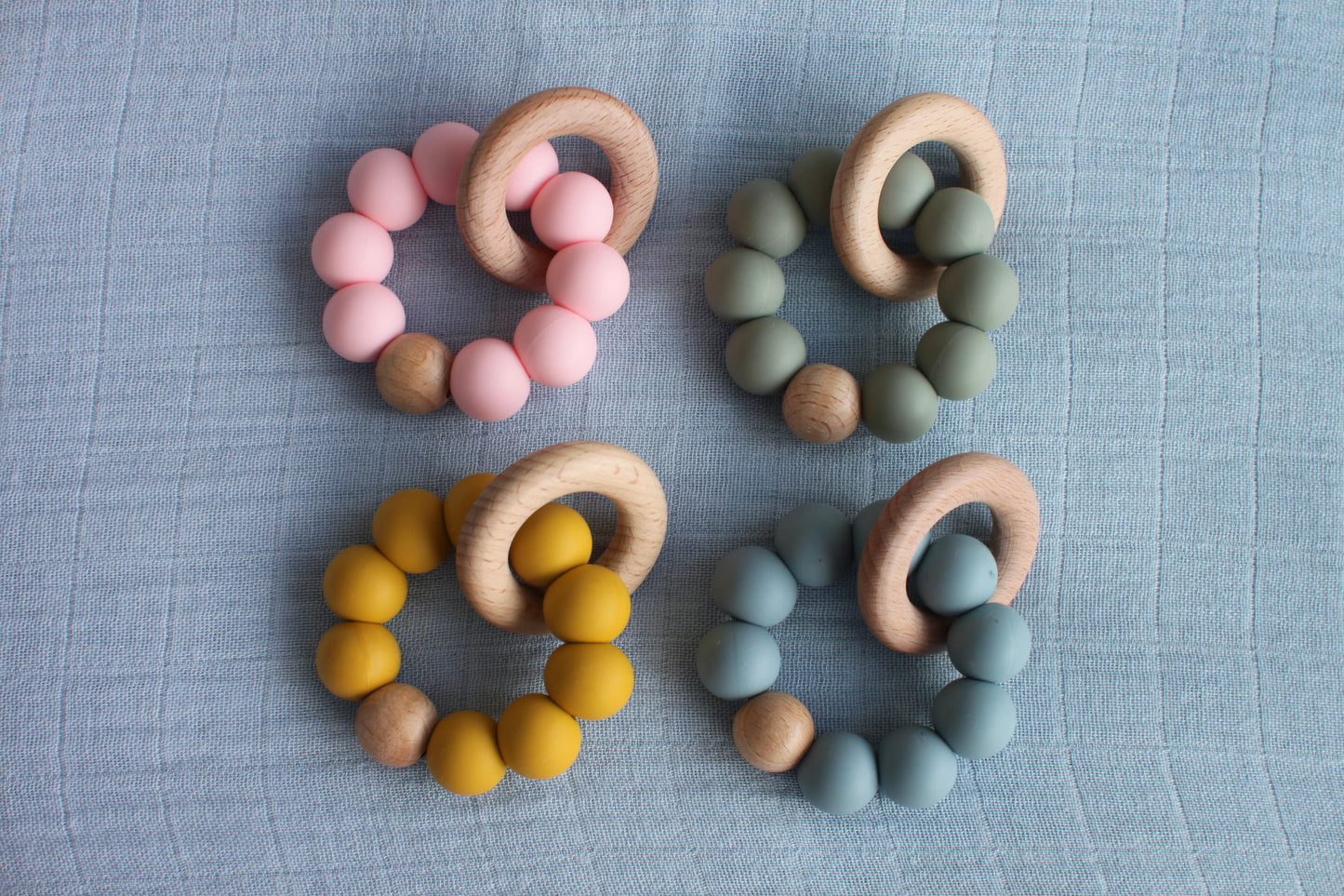 Four teethers made from silicone and beech wood are pictured. The colours of the beads are pink, green, blue and yellow (moving clockwise from the top left corner). 